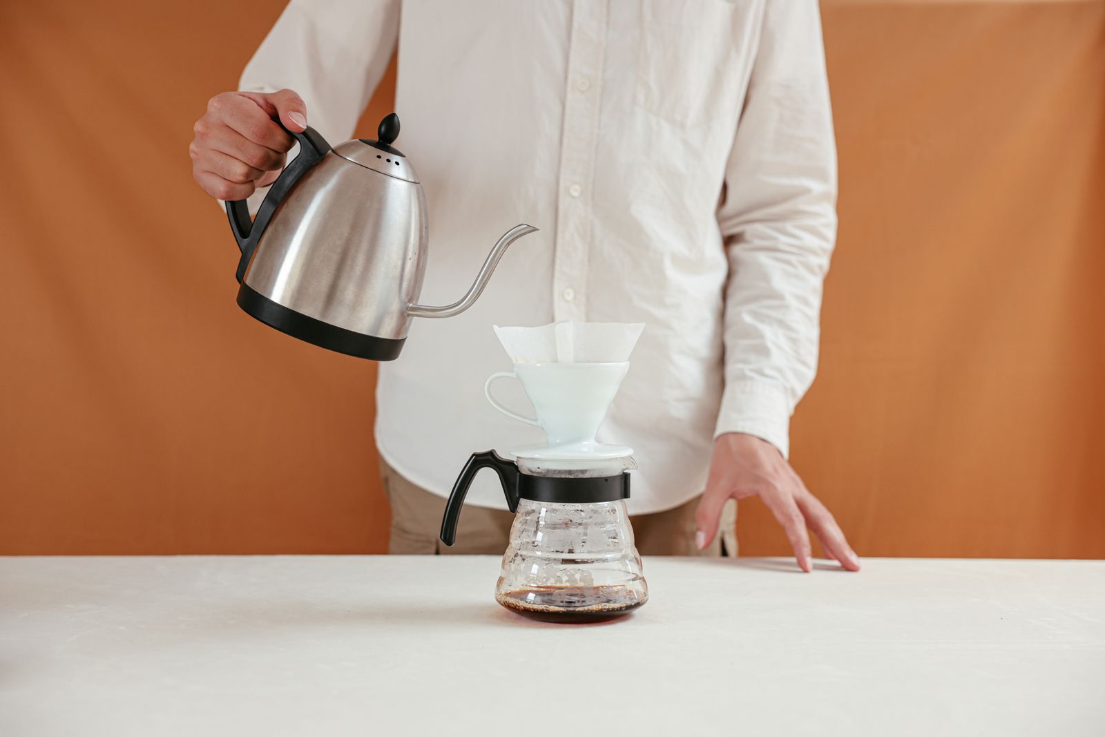 A gooseneck kettle pouring hot water into a white ceramic cone dripper set on a glass coffee server