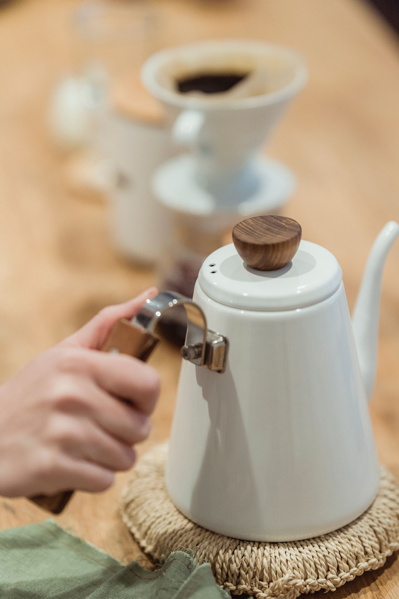 A white enamel gooseneck kettle held over a woven trivet, with a ceramic pour-over dripper in the background